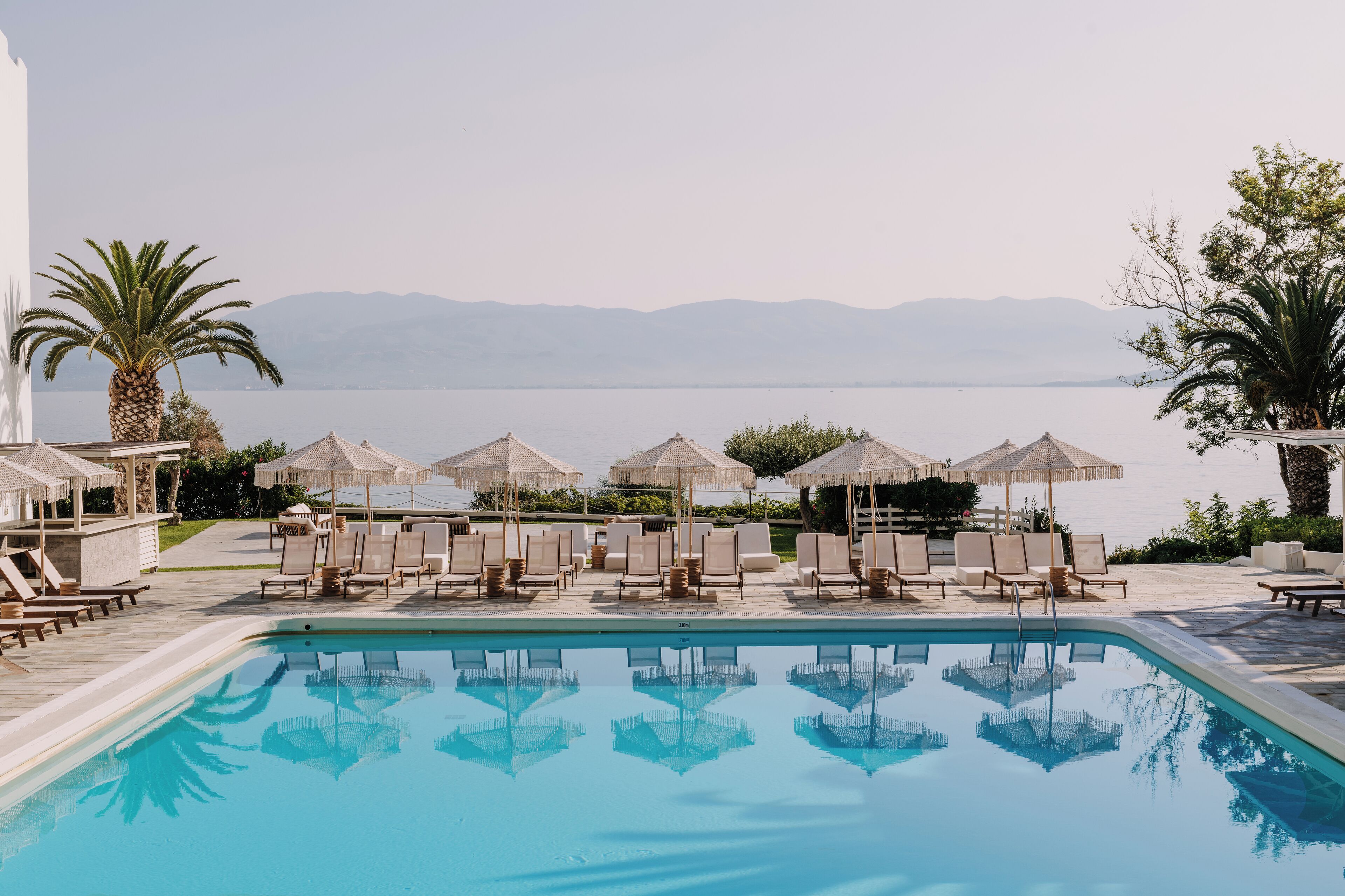 A poolside area with lounge chairs and umbrellas, a clear pool, and a view of the sea and mountains in the background.