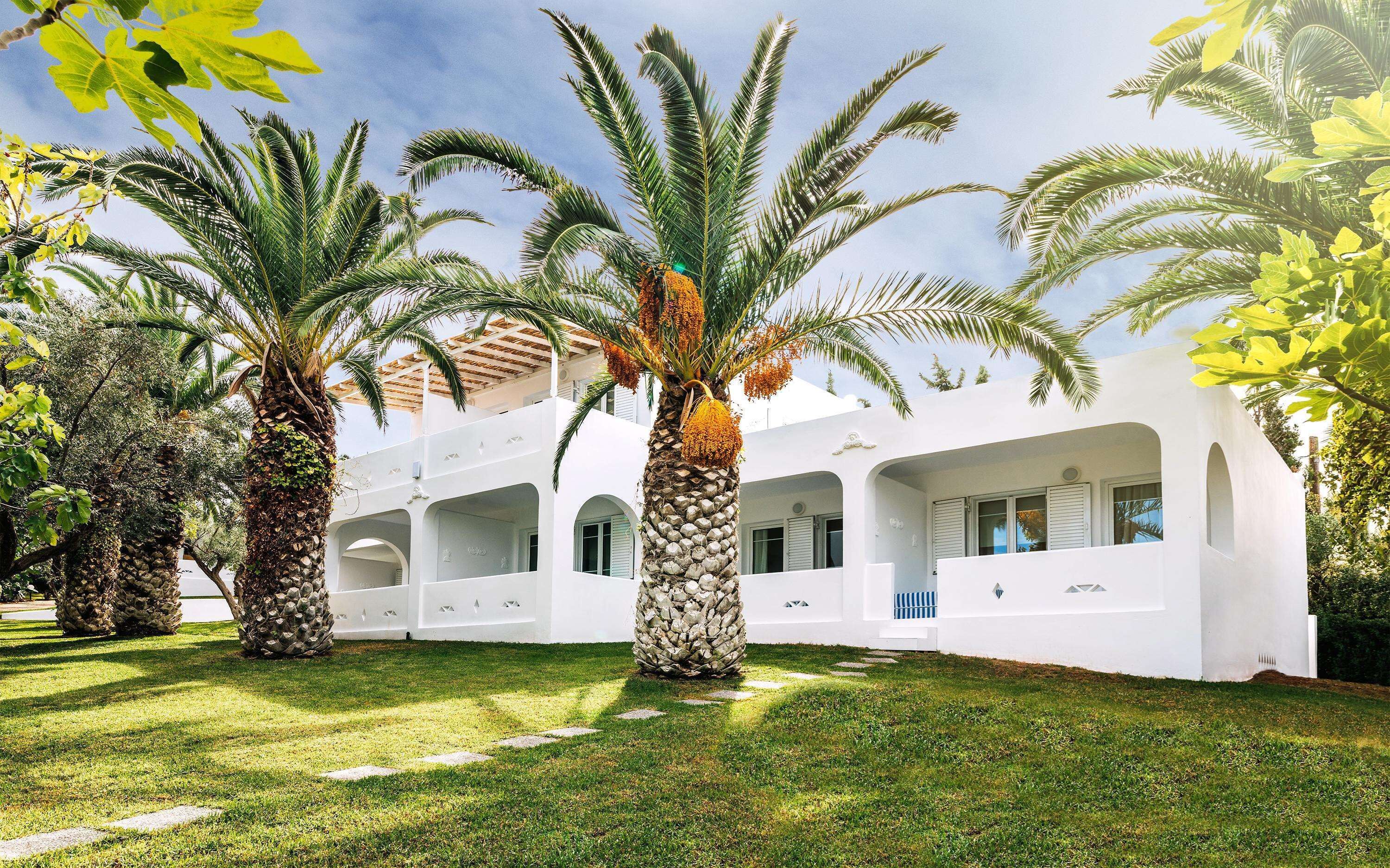 A white building with multiple balconies, surrounded by palm trees and a well-maintained lawn.