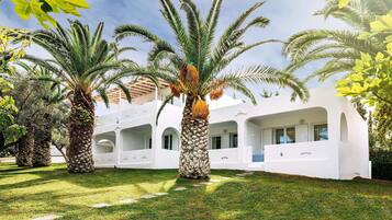 A white building with multiple balconies, surrounded by palm trees and a well-maintained lawn.