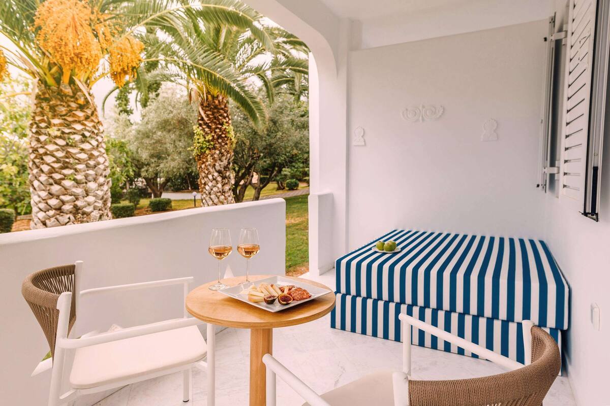 A balcony with a white chair, a wooden table with a plate of food and two glasses of wine, and a blue and white striped sofa.