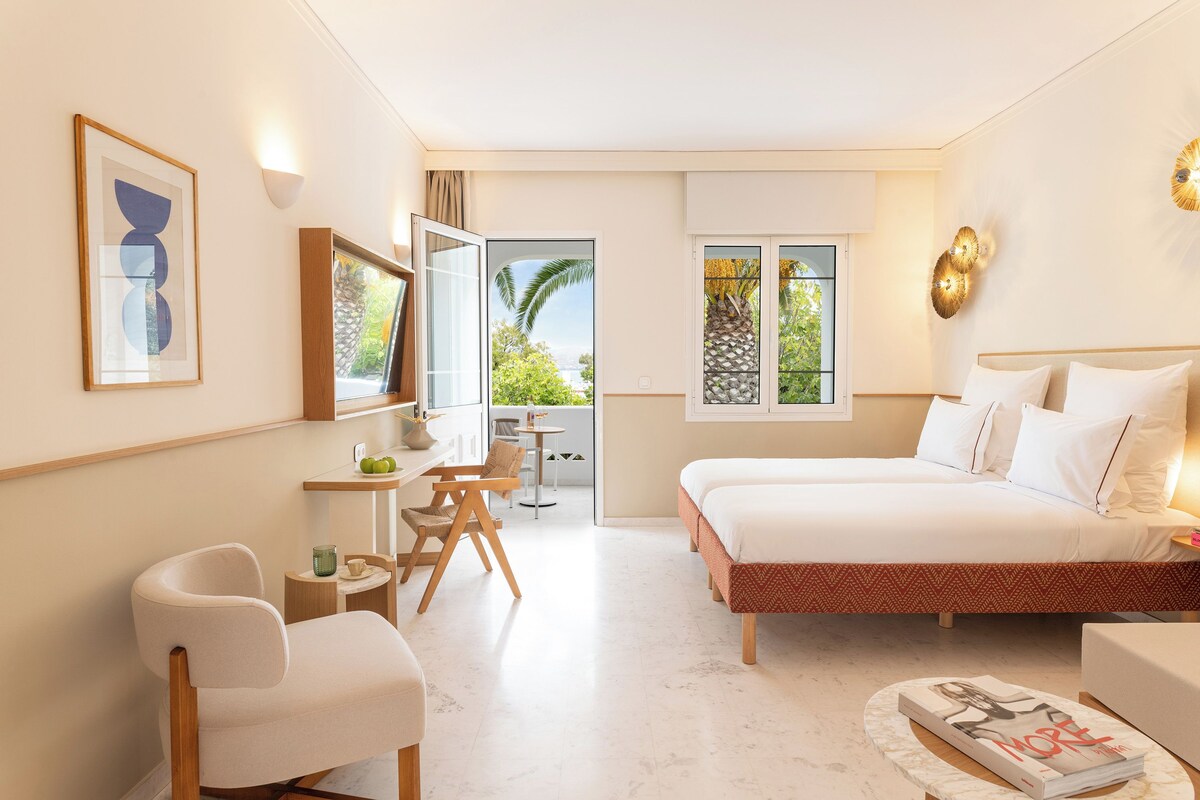 A modern hotel room with a bed, a desk, and a chair. There is a window with a view of greenery and a door leading to an outdoor area.