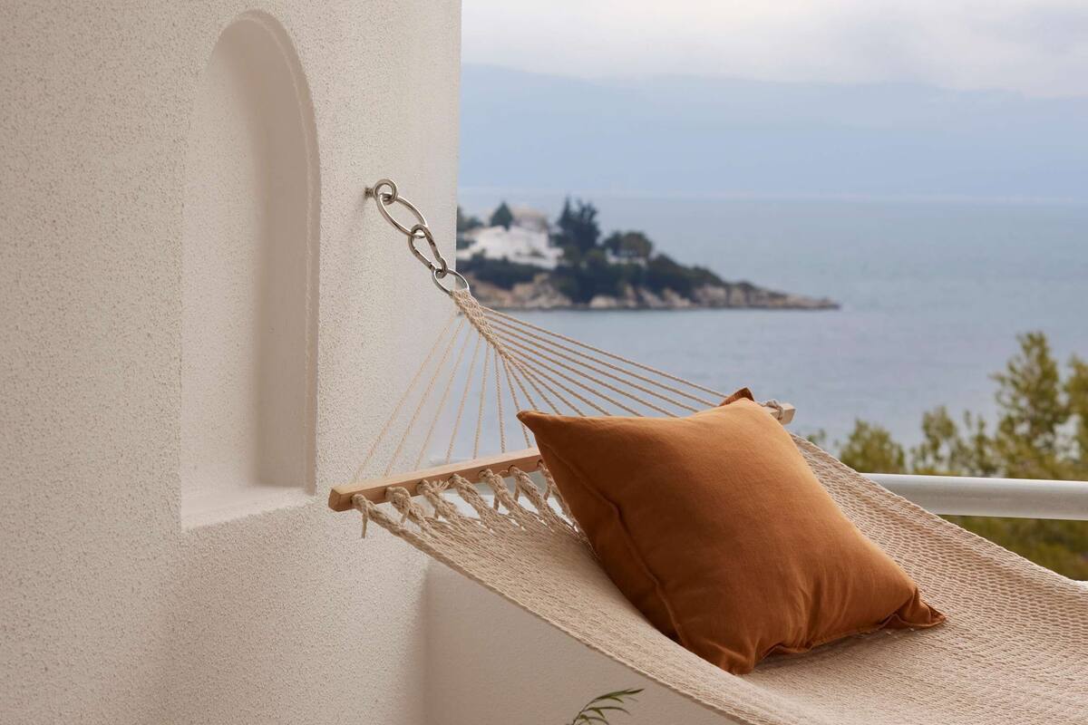 A hammock with a cushion, a bowl of apples, and a potted plant on a balcony with a view of the sea.