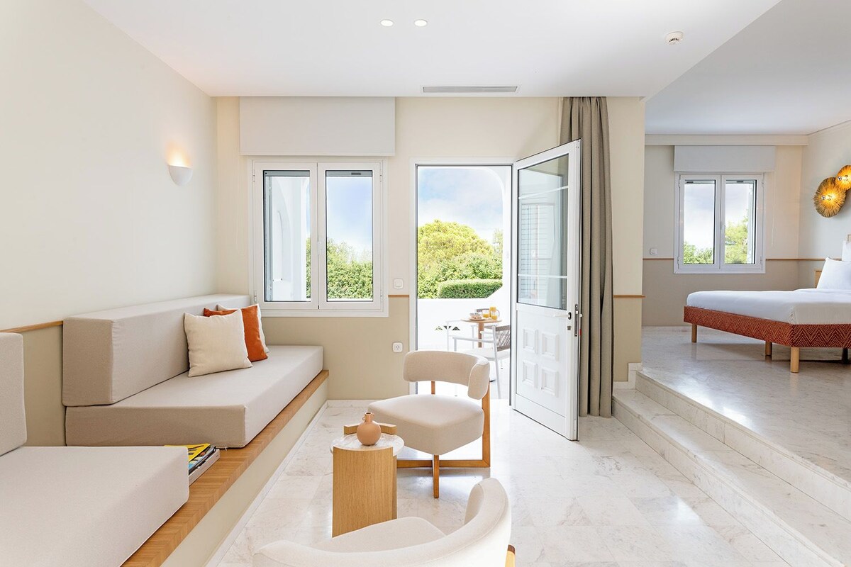 A modern hotel room with a sofa, a chair, a small table, and a bed. There are windows providing natural light and a view of greenery outside.