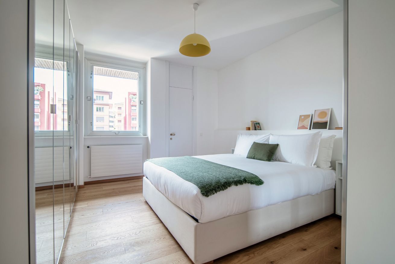 A bedroom with a large bed, a green throw blanket, white bedding, a yellow pendant light, and a view of buildings through a window.