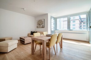 Ein modernes Wohnzimmer mit einem Holzesstisch, einem beigefarbenen Sofa und einem großen Fenster mit Blick auf die Stadt.