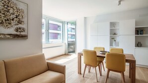 A modern living room with a sofa, dining table, and chairs. There is a large window with a view of buildings outside.