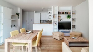 A modern living space with a wooden dining table, yellow chairs, a beige sofa, a TV, and built-in shelving with books and plants.