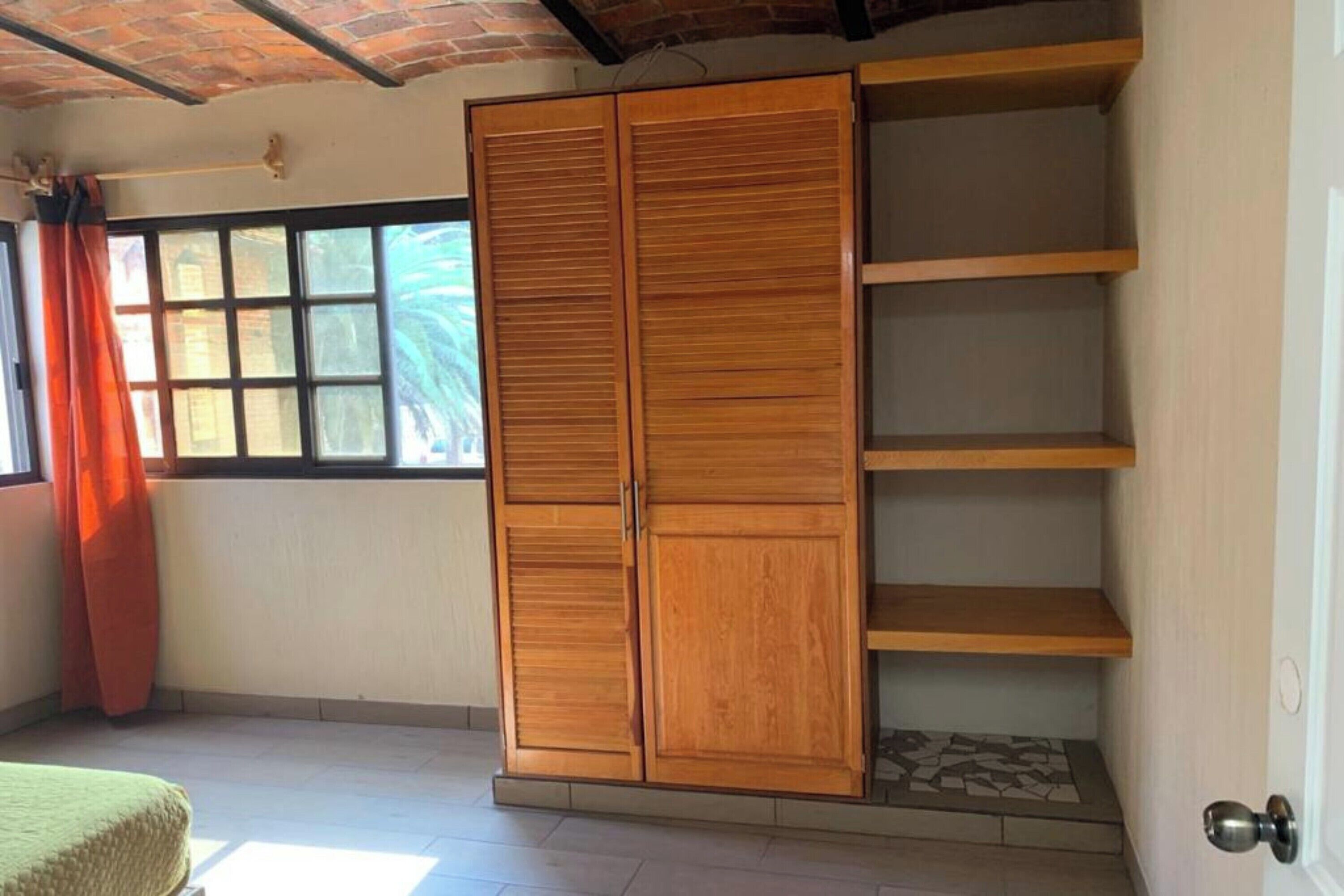 Una habitación con un amplio armario de madera, una ventana con cortinas naranjas y suelo de baldosas.