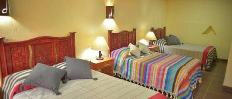 Habitación de hotel con tres camas, cada una con un edredón de estampado diferente. Se observan lámparas de noche y un reloj de pared.