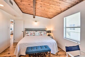 Een slaapkamer met een houten plafond, een groot bed met bloemenkussens, een bankje, een nachtkastje met een lamp en een raam met jaloezieën.