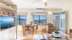 Una sala de estar moderna con comedor, ventanas amplias con vista al mar, un ventilador de techo y un televisor sobre un mueble.