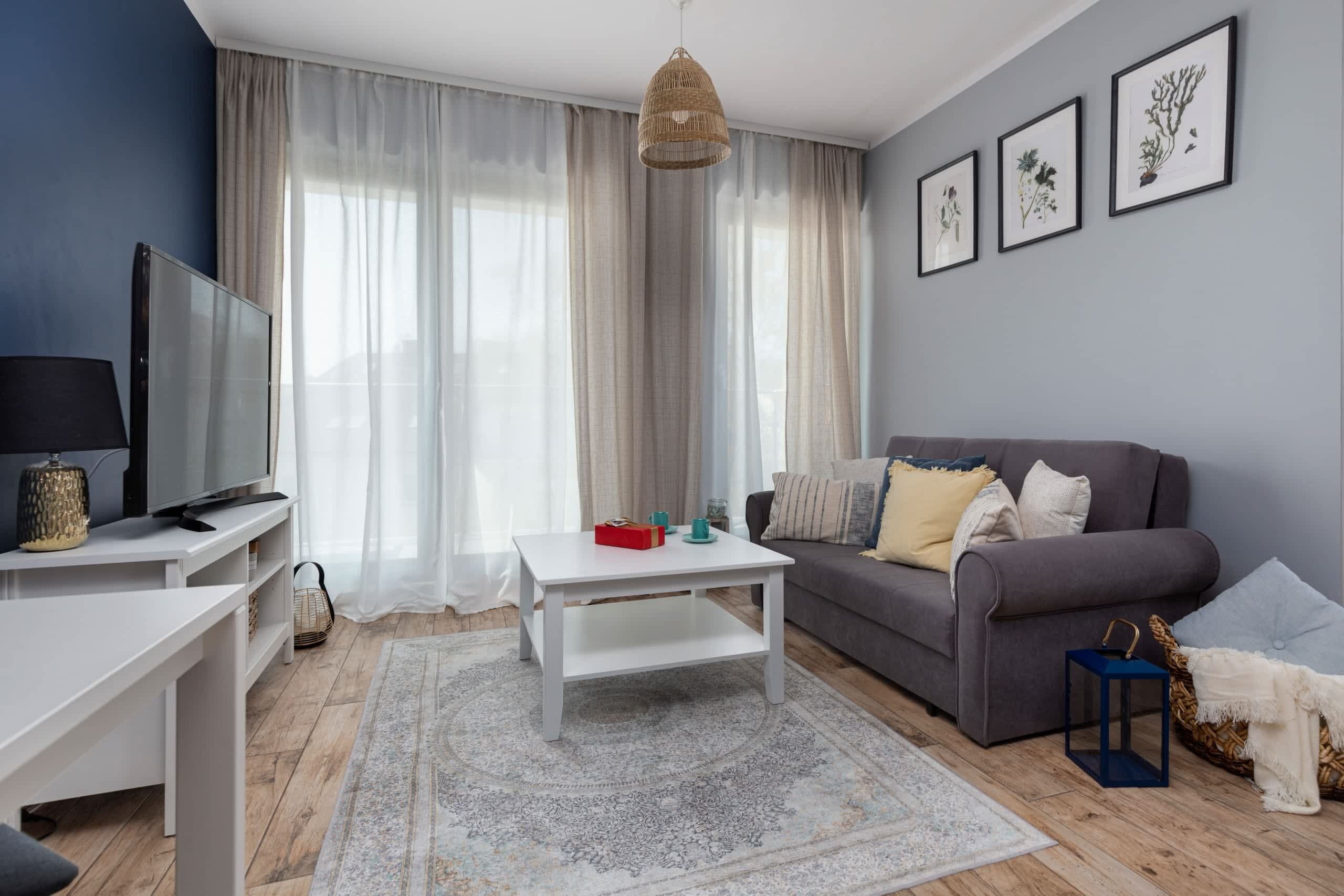 A living room with a TV, white coffee table, grey sofa, and a large window with sheer curtains.