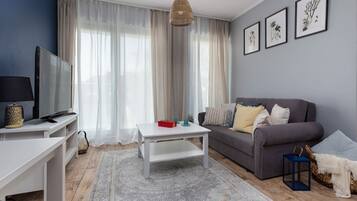 A living room with a TV, white coffee table, grey sofa, and a large window with sheer curtains.