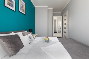 A neatly made bed with a tray of fruit and glasses, a nightstand with a lamp, and a teal wall with framed pictures.