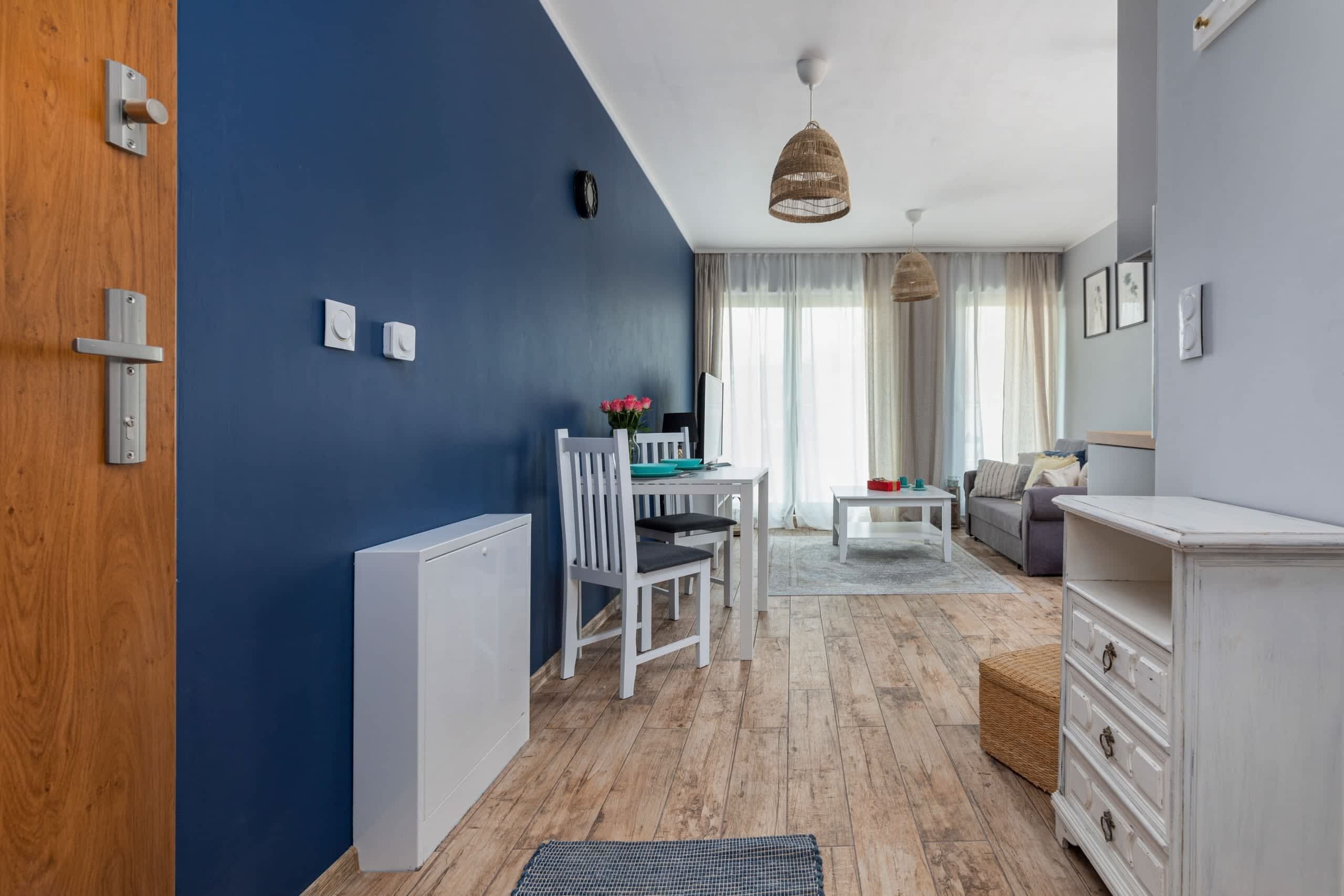 Een kamer met een blauwe wand, een houten deur, een witte radiator, een eettafel met stoelen, een bank, een witte dressoir en een hanglamp.