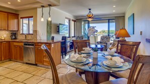 A modern kitchen with wooden cabinets, a dishwasher, and a coffee maker. There is a dining area with a glass table and chairs, and a view of the outdoors through large windows.