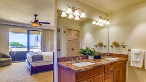 A hotel room with a balcony, a bed, a bathroom with double sinks, a shower, and a ceiling fan.