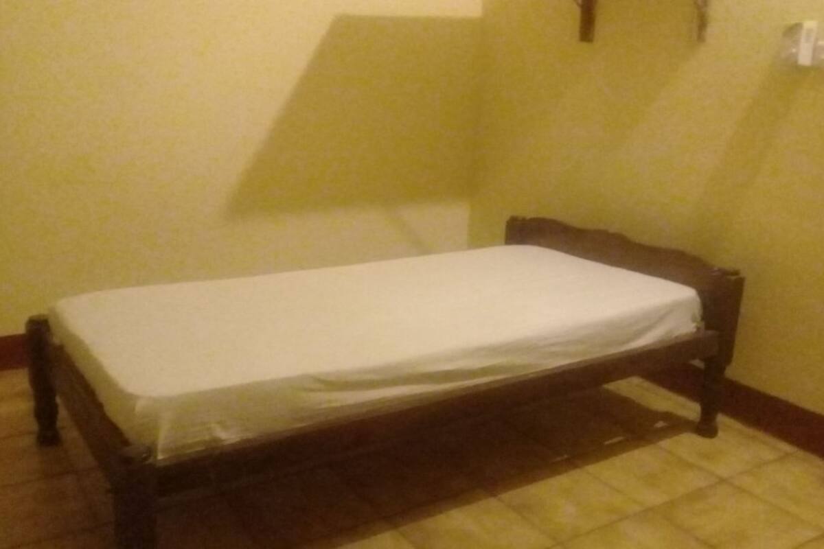 A small, single-bed room with a wooden bed frame and a white mattress. The room has a tiled floor and a plain yellow wall.