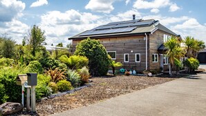 A log cabin with solar panels, surrounded by a variety of plants and a gravel driveway.
