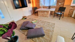 Un salon avec un téléviseur, un mur en briques, un coin repas avec des chaises et un petit îlot de cuisine.