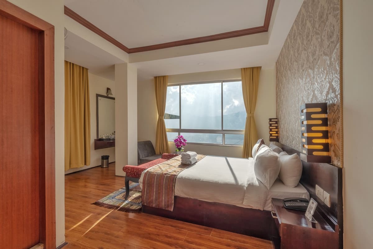 Habitación de hotel con piso de madera, una cama con sábanas blancas, una mesita de noche con teléfono y lámpara, una ventana con cortinas y una lámpara fijada a la pared.