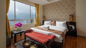 A hotel room with a large bed, a small table with a vase of flowers, a bench, and a view of the city through a large window.