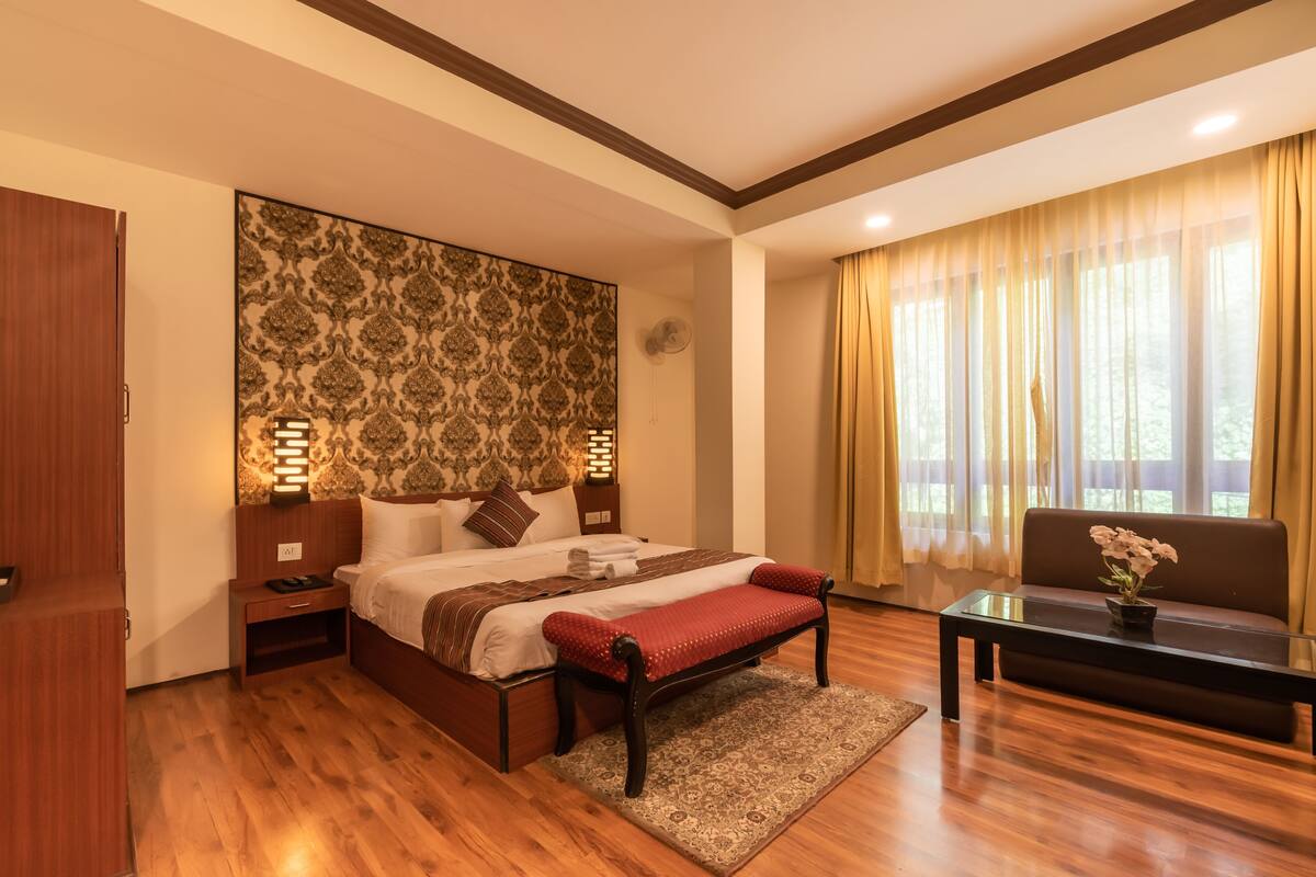 Una habitación de hotel con cama, mesitas de noche, un banco, un sofá, una mesa con flores, un reloj de pared y una ventana con cortinas.