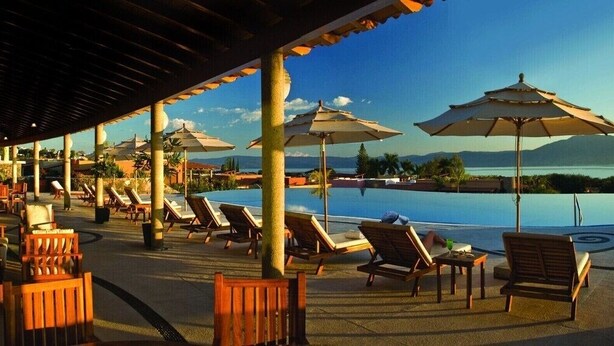 Zona junto a la piscina con sillones de descanso y sombrillas, con vistas a un lago y montañas.