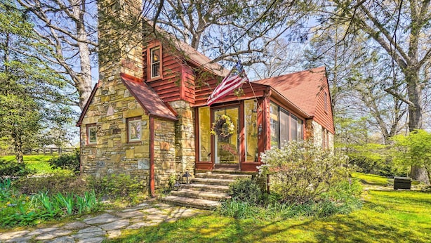 A stone house with a red roof and a porch, surrounded by lush greenery and trees. - Dreamy Homestead Escape: Patio, Ranch On-Site (Crossville)