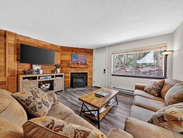 A cozy living room with a fireplace, wooden walls, a flat-screen TV, and a large window offering a view of the outdoors.