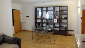 A modern living room with a glass-top dining table, clear chairs, a bookshelf, and a radiator. - Green view penthouse with beautiful terrace and playground (Lazio)