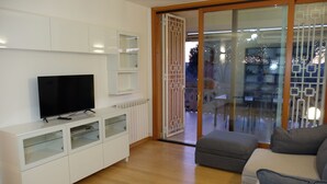 A modern living room with a flat-screen TV on a white cabinet, a grey sofa, and a glass door leading to an outdoor area. - Green view penthouse with beautiful terrace and playground (Lazio)
