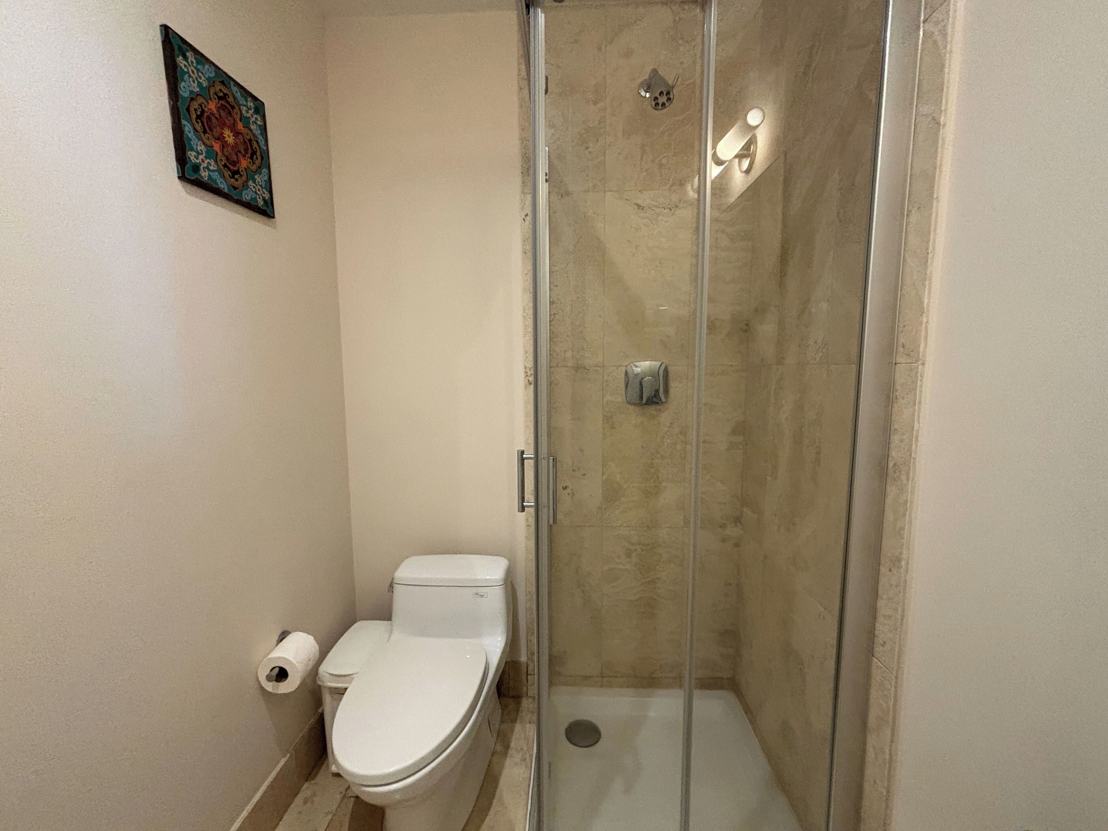 A bathroom with a glass-enclosed shower, a toilet, and a roll of toilet paper on the floor.