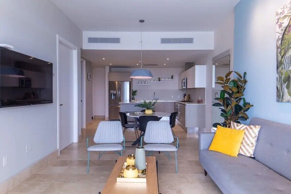 A modern living room with a grey sofa, a dining table with chairs, and a kitchen in the background.