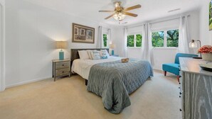 Quarto com cama, mesas de cabeceira, escrivaninha, cadeira, ventoinha no teto e janelas com cortinas.