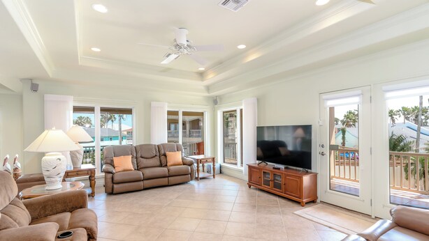 A spacious living room with a ceiling fan, a flat-screen TV, and a balcony with a view of palm trees.