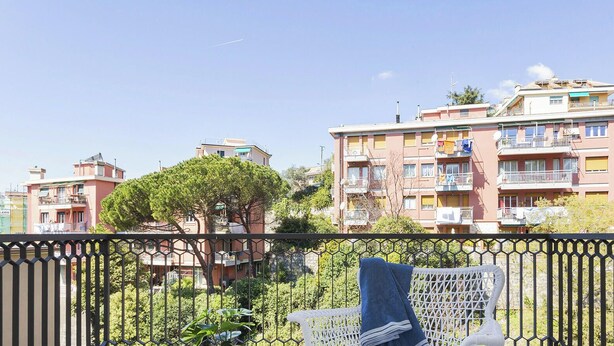 A balcony with a white chair, a small table, and a potted plant. In the background, there are multi-story buildings with balconies. - Shelley Apartments - Tree by Wonderful Italy (Genova)