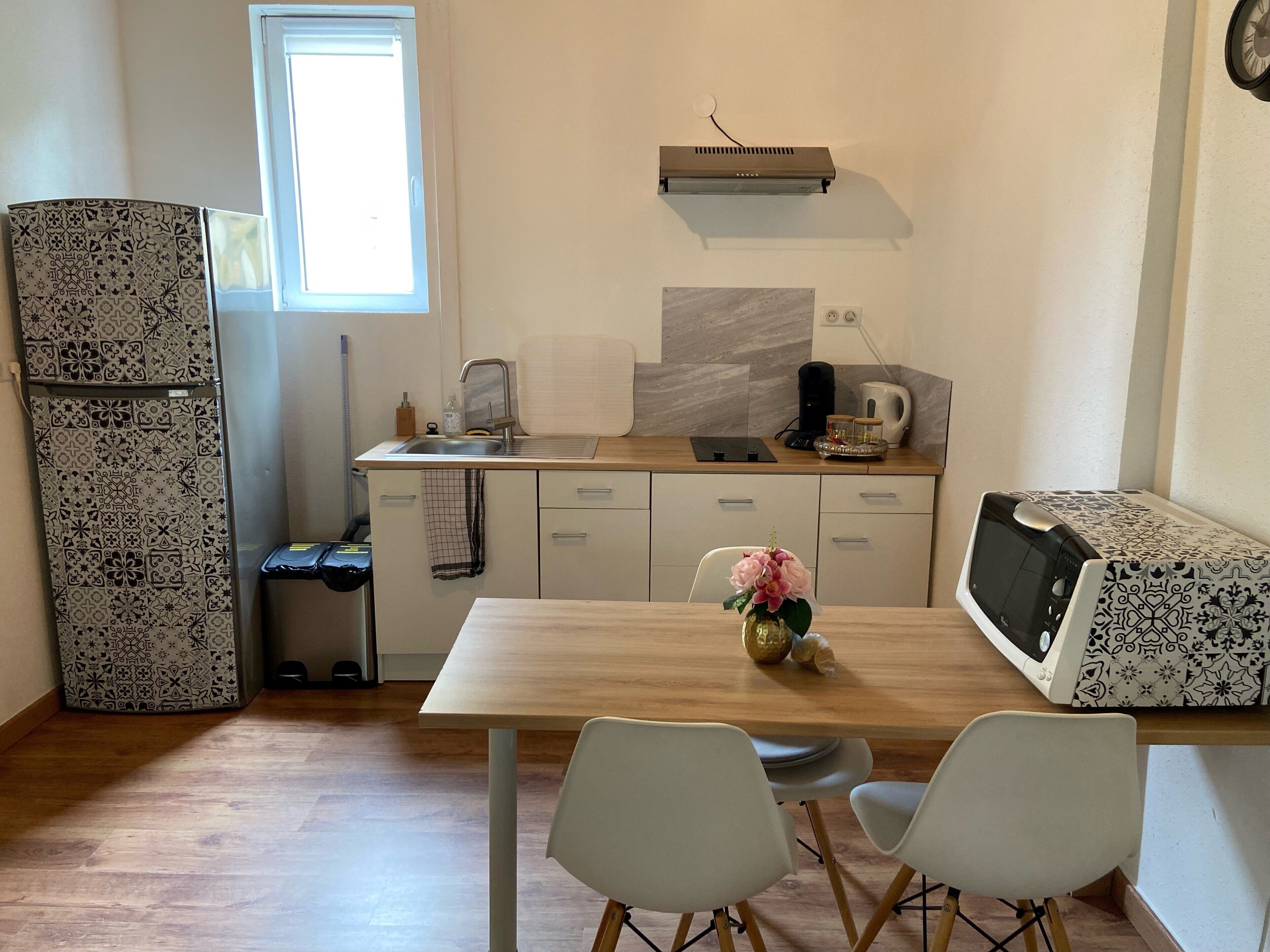 A compact kitchen with a sink, a microwave, and a coffee maker. There is a window providing natural light, and a refrigerator with patterned doors.