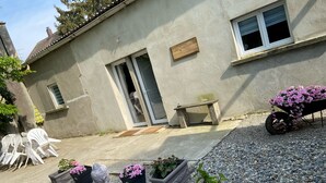 Una casa con fachada de concreto, una puerta y una ventana. Hay plantas en macetas con flores rosas, un banco y un carretón con flores.