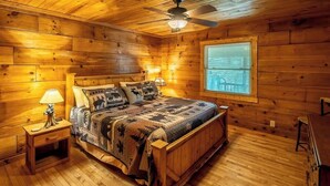Een slaapkamer in een houten hutje, met een bed, nachtkastjes, een plafondventilator en een raam met jaloezieën.