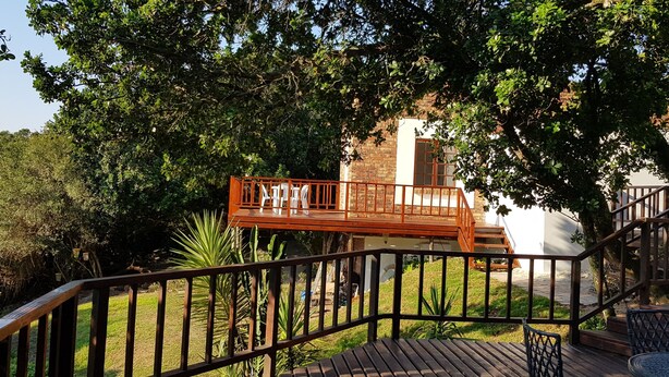 A wooden deck with a railing, surrounded by greenery and a brick building in the background. - Country Cottage in Conservation Area off Pari Park (Gqeberha)