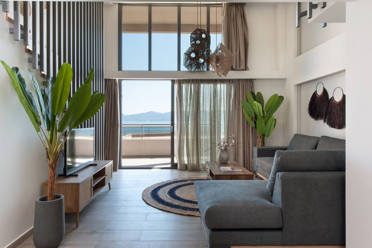 A modern living room with a grey sofa, a wooden TV stand, a large potted plant, and a view of the sea through a sliding glass door.