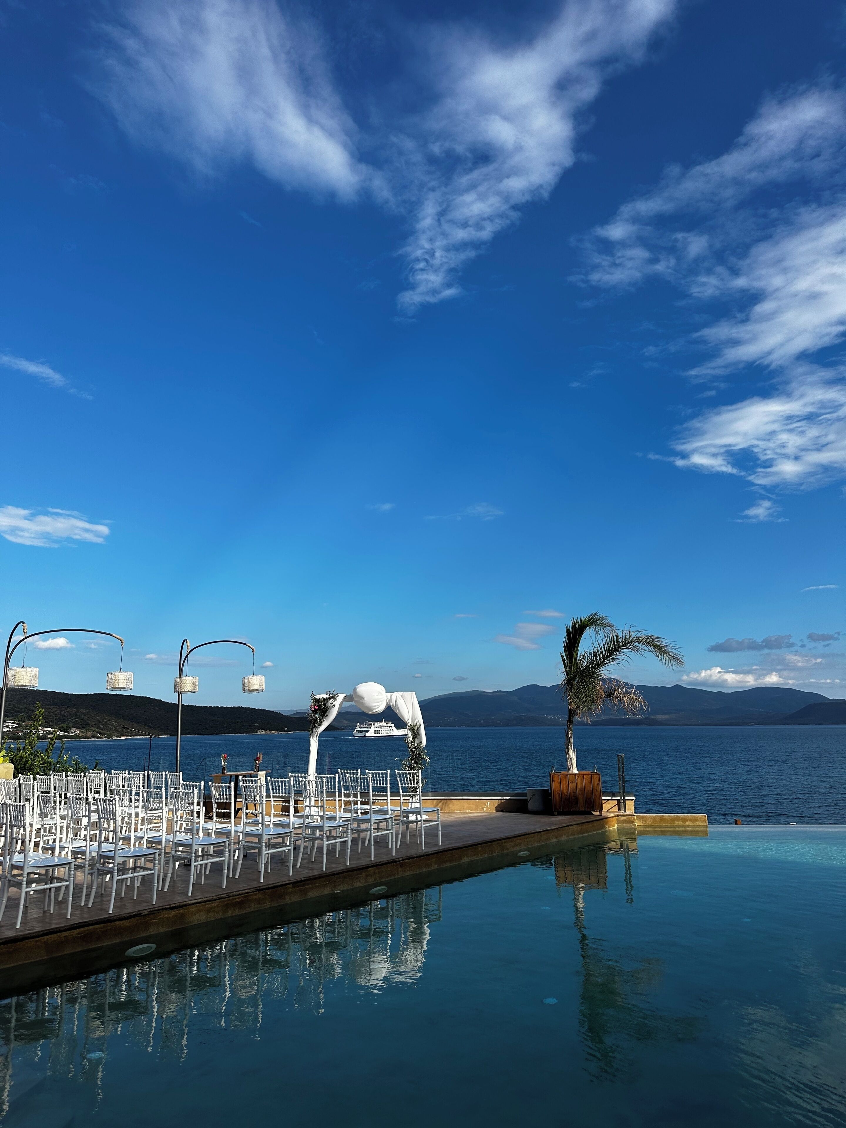 An outdoor pool area with white chairs and a white arch structure, overlooking a body of water and distant mountains.