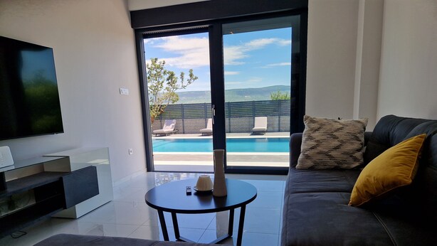 A modern living room with a large TV, a sofa, and a small table. There is a view of a pool and outdoor furniture through a glass door. - Luxury villa with private swimming pool (Zmijavci)