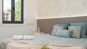 Un lit bien fait, avec une couette à motifs floraux, une assiette de fruits et une table de chevet sur laquelle se trouvent une lampe et un objet décoratif.