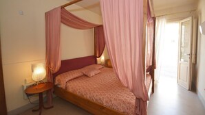 Uma cama com dossel e cortinas cor-de-rosa, uma mesinha de cabeceira com abajur e vista para o exterior através de uma porta.