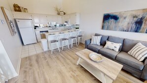 Eine moderne Küche mit weißen Schränken und einem Essbereich mit weißen Stühlen. Ein graues Sofa mit gestreiften Kissen und ein Couchtisch mit einem gewebten Korb sind ebenfalls vorhanden.