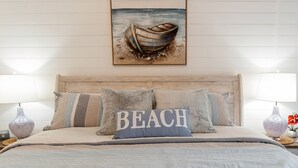 Un dormitorio con cabecera de madera, dos lámparas de noche, una cama con manta a rayas beige y azul, y un cojín con la palabra 'BEACH'.