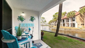 Un porche con sillas turquesas y una planta en maceta, con vista a un canal y una hilera de edificios con una palmera.
