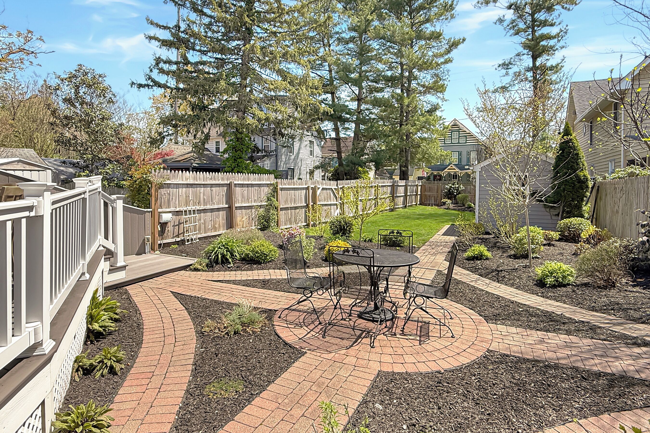 Une terrasse à l’arrière de la maison, avec une table et des chaises, entourée d’un chemin en briques et d’un jardin aménagé.